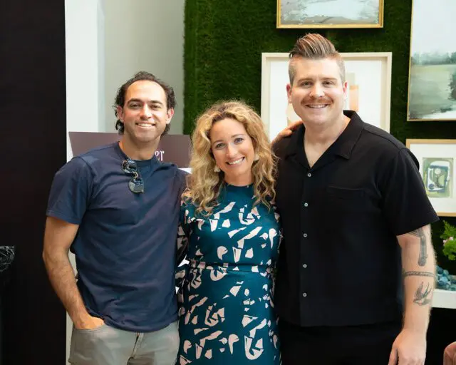 Three people smiling, two men and one woman, in front of a green wall with artwork.