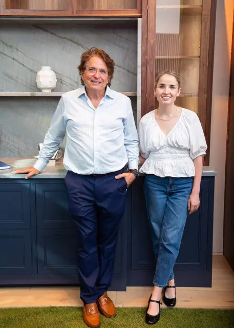 Man in blue checkered shirt and woman in white blouse stand in front of dark blue cabinets.