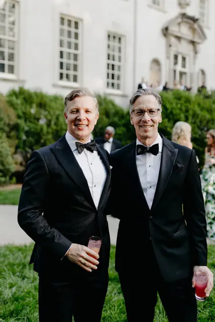 Two men in tuxedos holding pink drinks outside a white building.