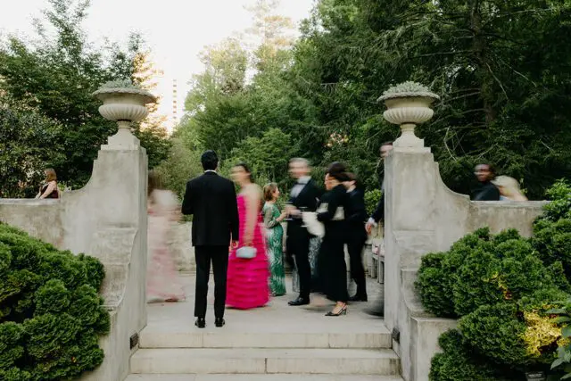 People in formal attire mingling outdoors near stone architecture and lush greenery.