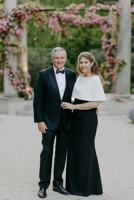 Man in tuxedo and woman in black dress with white cape stand outdoors