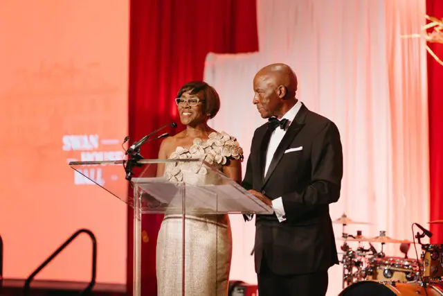 Woman in gold dress and man in tuxedo at podium with "Swan House Ball" on screen