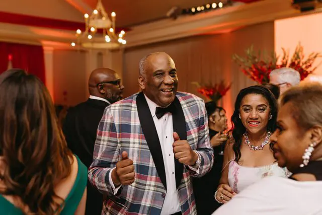 Man in plaid jacket and bow tie smiles, giving thumbs up.