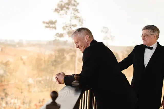 Two men in tuxedos stand on a balcony, one laughing and leaning on the railing.