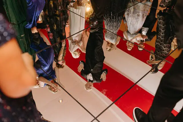 People in formal attire reflected in a mirrored ceiling above a red and white striped floor.