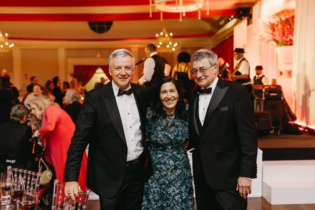 Three people in formal wear posing for a photo at an event with a band playing in the background.