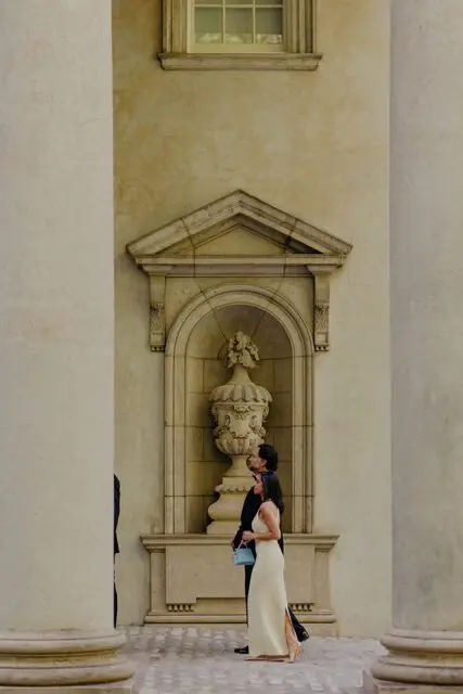 Couple in formal wear walking past a stone urn niche and columns