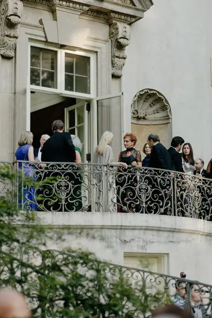 People in formal attire on a balcony of a stone building with ornate railings.