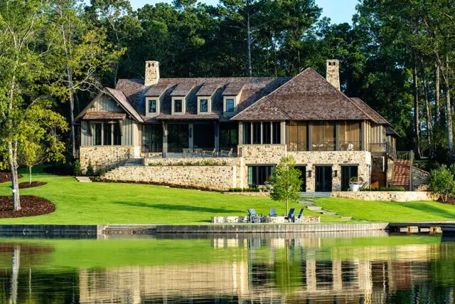 Stone and wood home with multiple gables and dormer windows on a grassy lawn by a lake.