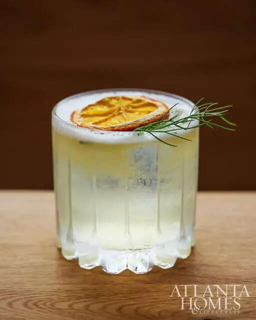 Cocktail with frothy top, garnished with dried orange slice and rosemary sprig.