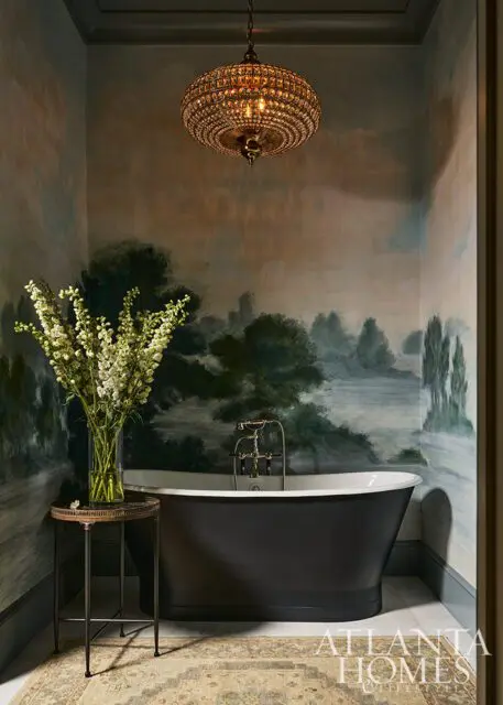 Dark freestanding tub with mural wallpaper, ornate chandelier, and floral arrangement.