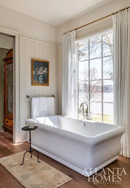White freestanding bathtub with a large window overlooking a lake, paneled walls, and a floral