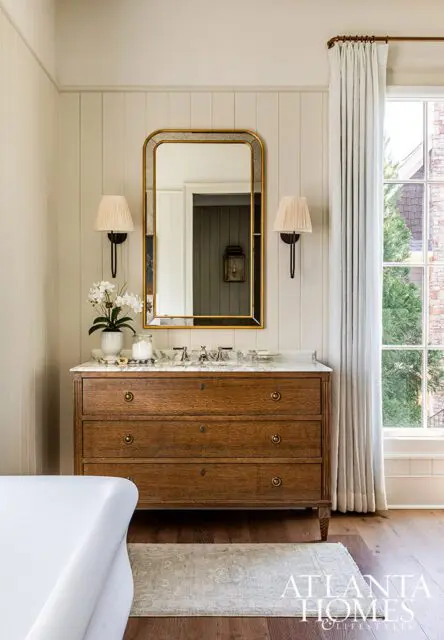 Wood vanity with marble top, gold mirror, and sconces. White bathtub in foreground.