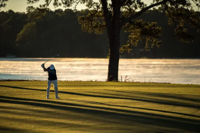 Golfer swings club on a green course with a lake and trees in the background at sunrise.