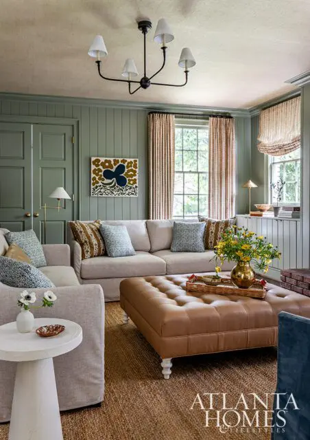 Living room with green paneled walls, neutral sofas, tufted leather ottoman, and floral curtains.