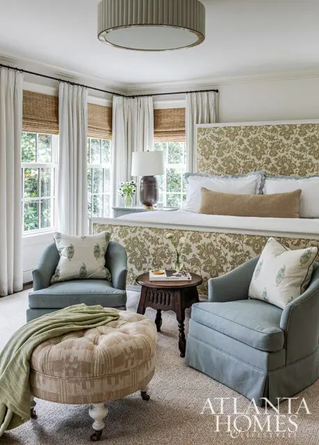 Bedroom with upholstered headboard, two blue armchairs, tufted ottoman, and patterned wallpaper.