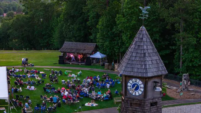 Outdoor concert with audience on lawn near wooden clock tower and stage.