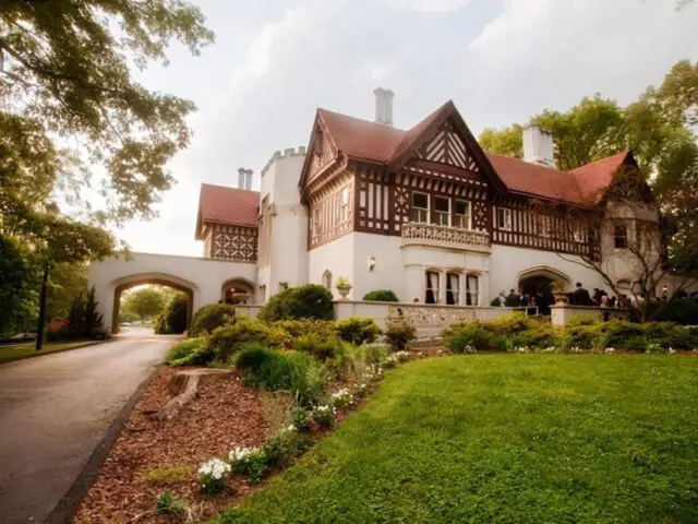 Tudor-style mansion with red tile roof and white stucco walls, surrounded by lush greenery.
