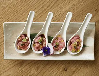 Four white ceramic spoons holding raw meat tartare with garnishes, on a rectangular platter