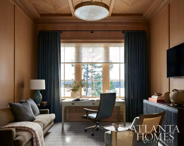Office with wood paneled walls, desk, chair, sofa, and window overlooking water and trees.