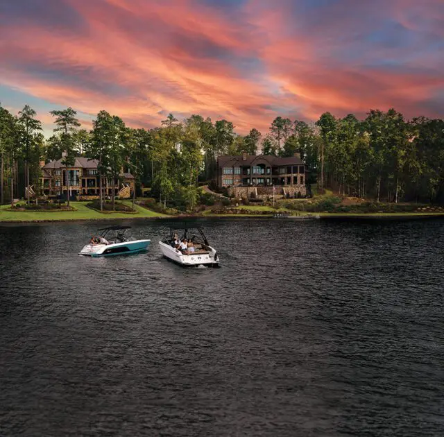 Two boats on a lake with luxury homes and trees in the background under a colorful sunset sky.