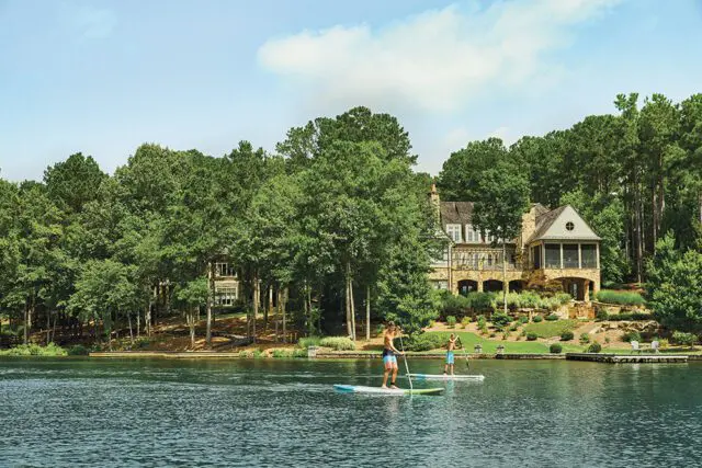 Two men paddleboarding on a lake in front of a large stone house surrounded by trees.