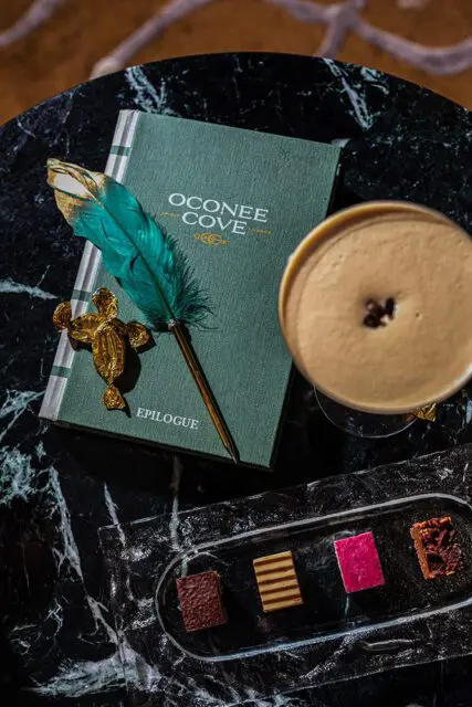 Green book with gold text 'Oconee Cove Epilogue', feather pen, and chocolates