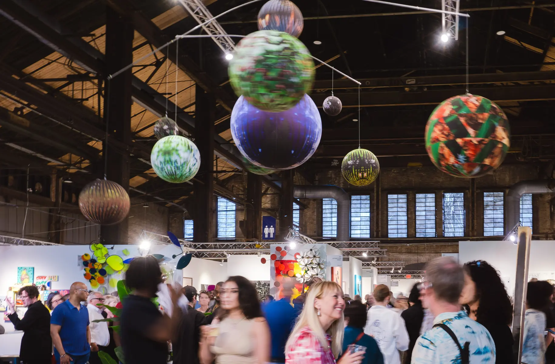 People at an art exhibition with large spherical art installations hanging from the ceiling.