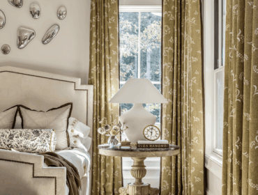 Bedroom with upholstered headboard, patterned curtains, and a round side table with a lamp and