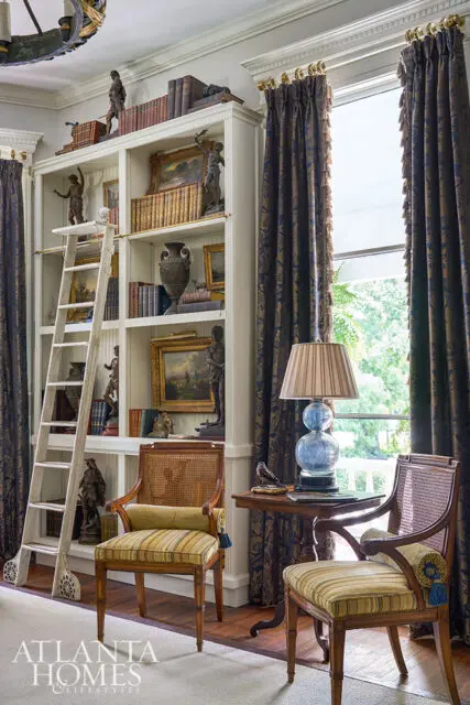 Built-in bookshelf with ladder, two chairs, and a lamp by the window.