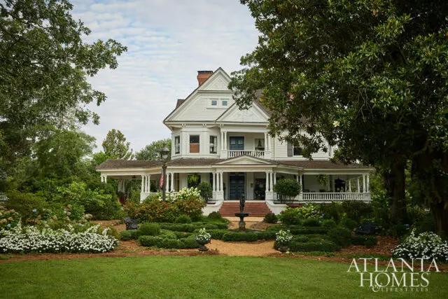 White Victorian house with wraparound porch, surrounded by lush gardens and trees.