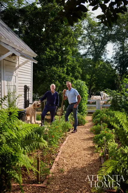 Two men and a dog stand on a gravel path in a garden with horses in the background.