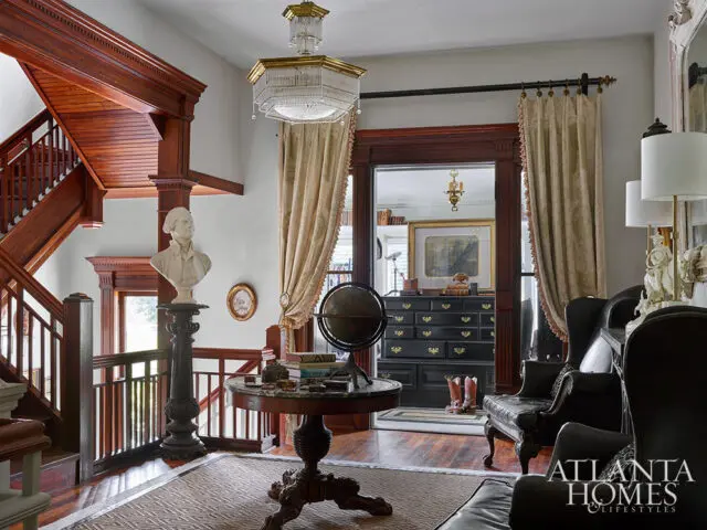 Foyer with ornate wooden staircase, bust on pedestal, globe on table, and dark leather chairs.