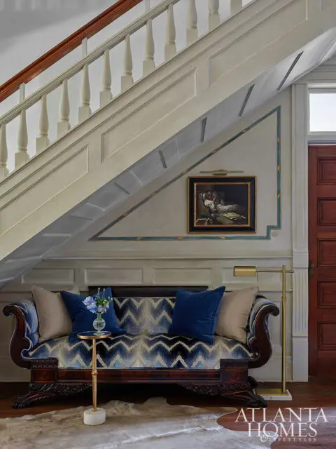Blue patterned sofa with dark wood frame under a staircase with a painting on the wall