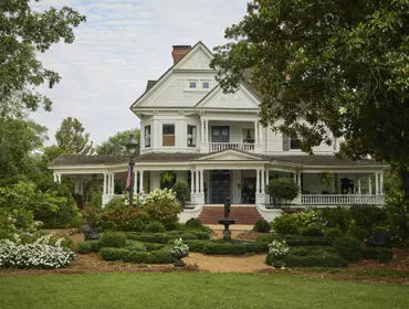 White Victorian house with wraparound porch, formal gardens, and fountain.