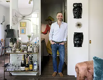 Man in blue shirt and jeans standing in hallway with bar cart and art on wall.