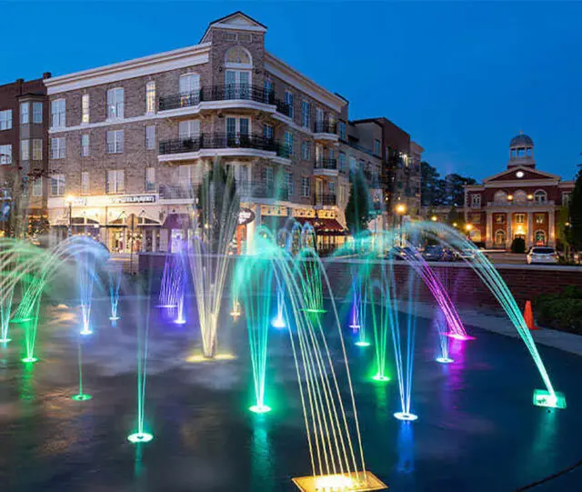 Colorful illuminated fountains in a plaza at dusk with brick buildings in the background.