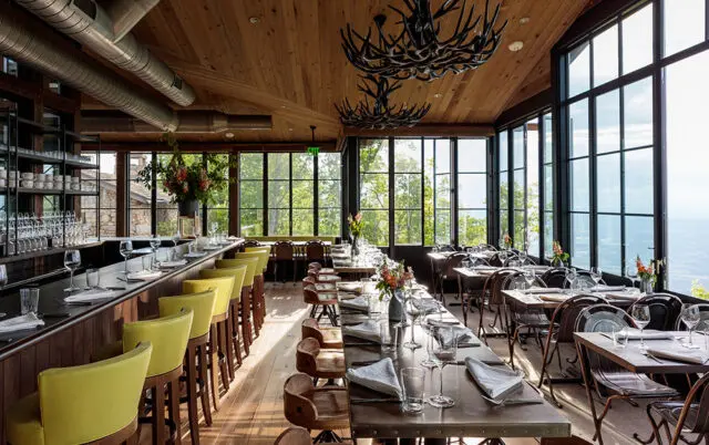 Restaurant dining room with bar, tables set for dinner, and large windows overlooking a view.