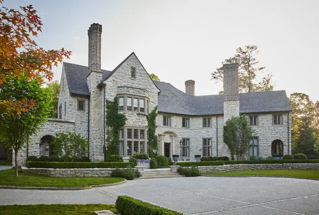 Stone mansion with multiple chimneys, arched entryway, and manicured hedges.