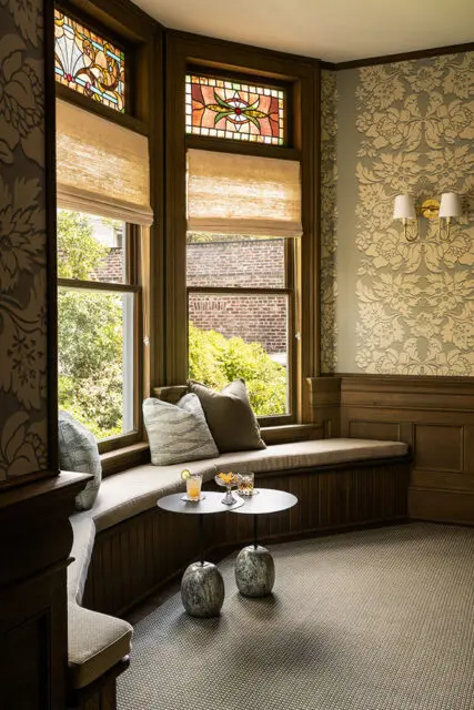 Window seat with stained glass, patterned wallpaper, and two small tables with drinks.