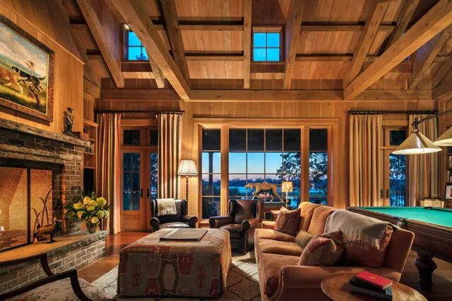 Living room with wood paneled walls, exposed beams, stone fireplace, and large windows overlooking