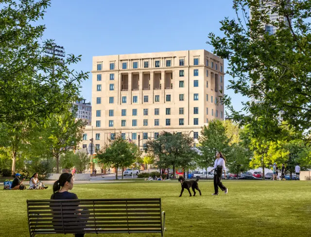 People and a dog in a park with a light-colored building in the background.