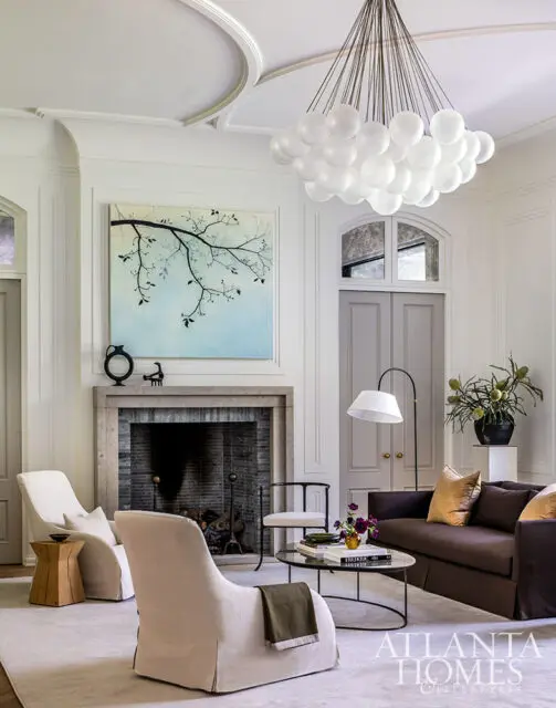 Living room with fireplace, brown sofa, and white armchairs. Large bubble chandelier hangs overhead.