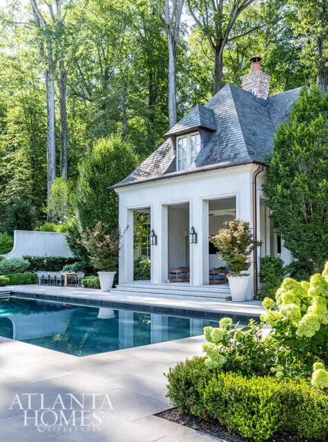 Poolside cabana with arched openings and slate roof, surrounded by lush landscaping and trees.