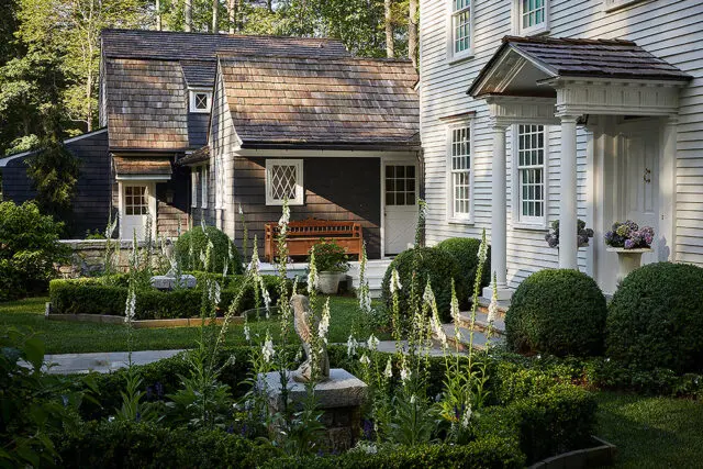 Shingled house with white columns and porch, surrounded by manicured gardens and foxglove flowers.