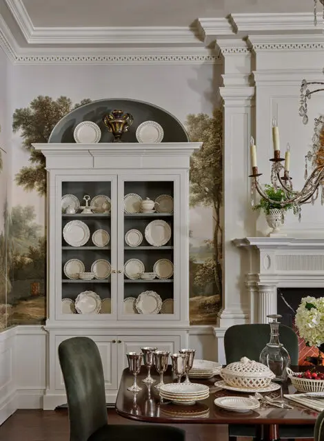 Dining room with a white china cabinet filled with white dishes, landscape wallpaper, and a table