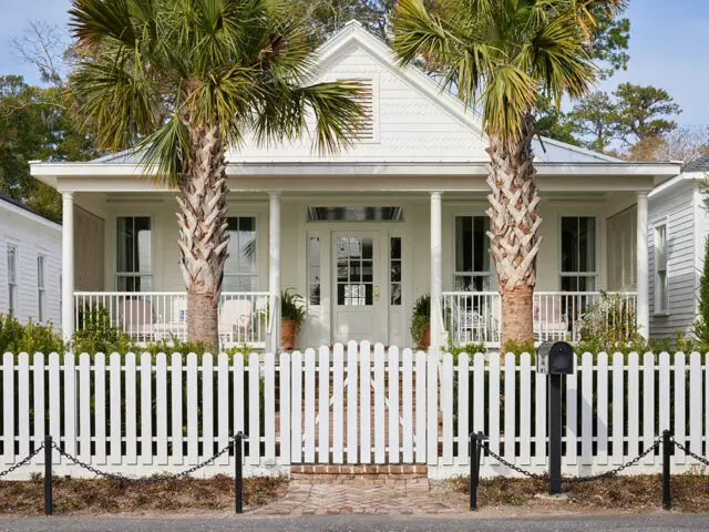 White house with a porch, palm trees, and a white picket fence.