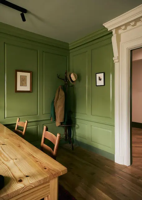 Green paneled room with wooden table, chairs, coat rack with hat, and framed art.