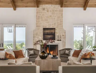 Living room with stone fireplace, wood beams, and neutral sofas with orange accents.