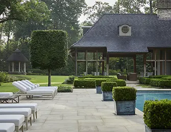 Stone patio with lounge chairs and pool next to a house with dark roof and dormer window.